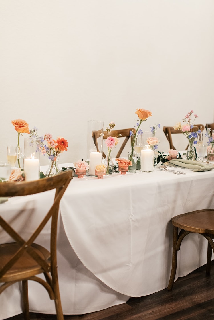 Long reception table set with orange roses, greenery, and glowing candles between empty cross-back chairs.