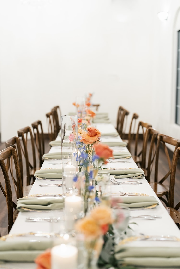 A long reception table set with sage napkins, gold-rimmed chargers and candles, orange and blue floral arrangements running down the center in a softly lit room.