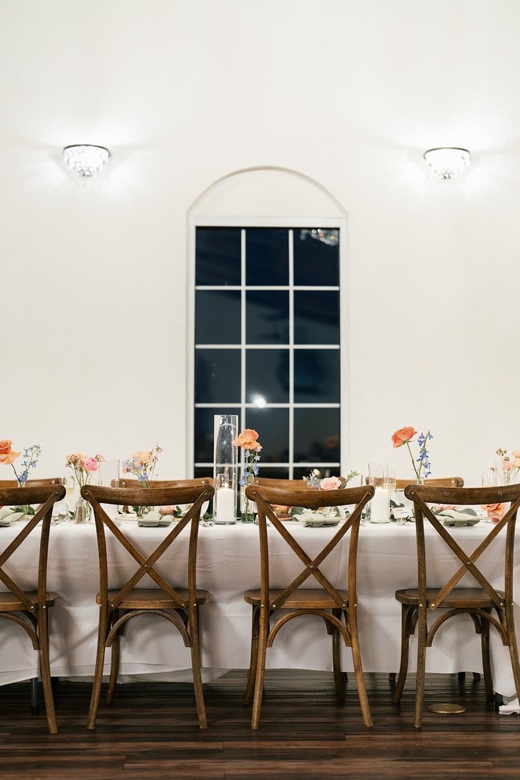 Row of empty wooden chairs facing a white-linened head table decorated with orange and pink florals beneath an arched window.