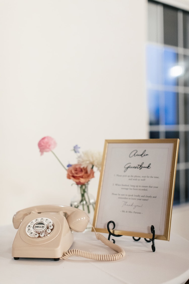 Vintage cream rotary phone beside a framed 'Audio Guestbook' sign and a small vase of pink and orange flowers on a reception table.