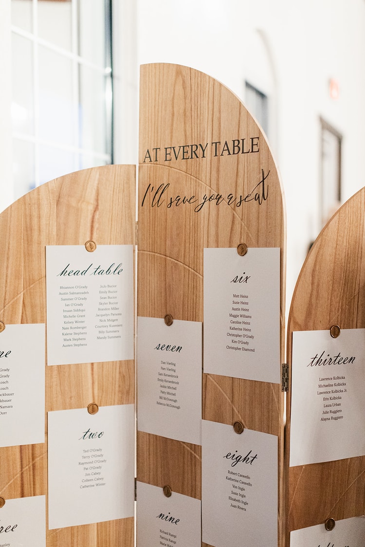 Close-up of a wooden seating chart with tables labeled 'six,' 'seven,' 'eight,' and 'nine' listing guest names in calligraphy.
