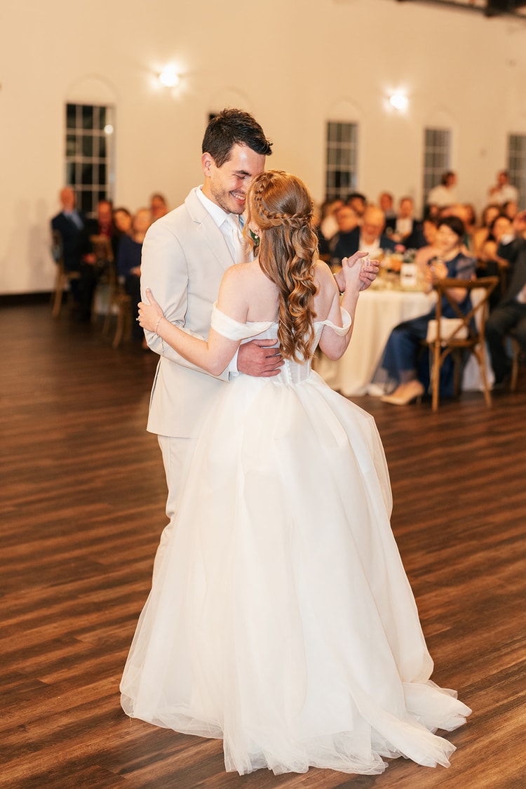 Groom embraces the bride from behind during their first dance, her braided half-up hairstyle catching the reception lights.