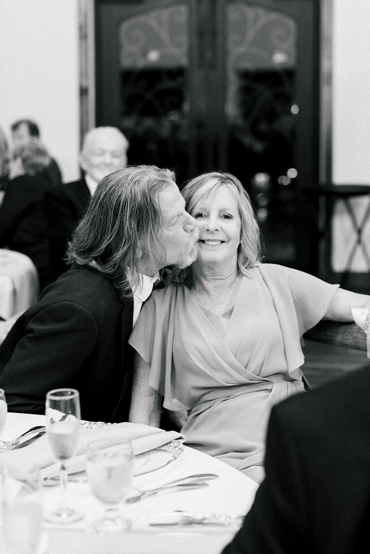 Black and white candid of an older man kissing a woman's cheek as she smiles at the reception table, other guests seated nearby.