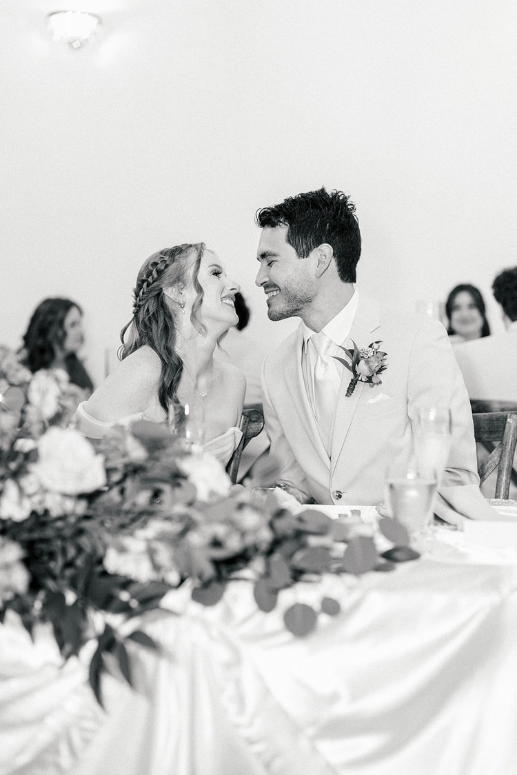 Black and white close-up of bride and groom laughing together at the head table, a floral garland draped along the tablecloth in front of them.