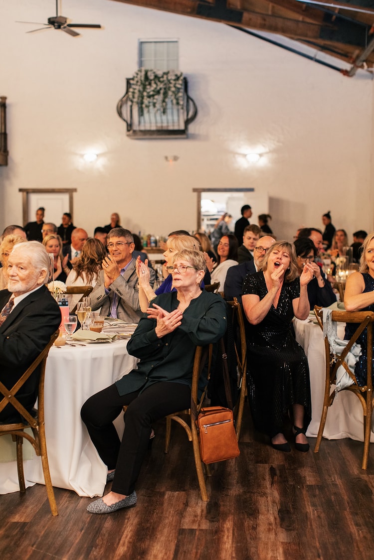 Wedding guests seated at reception tables applaud and cheer, one woman laughing mid-clap beneath string lights and open doorways.