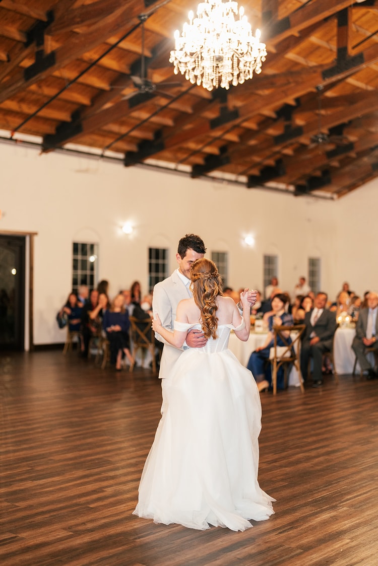 Bride and groom dance close together beneath a crystal chandelier as guests look on from surrounding tables.