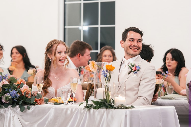 Bride and groom laugh at the head table during a reception toast, her braided hair and his boutonniere lit by warm string lights.
