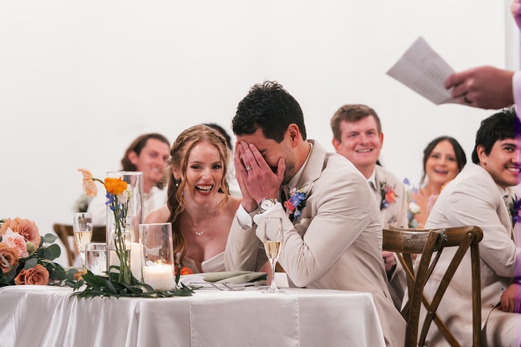Groom covers his face laughing as the bride beams beside him at the head table, guests behind them smiling during toasts.