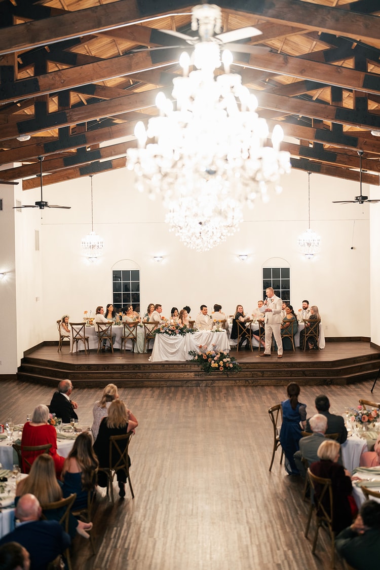 Wide view from above the dance floor showing the head table on stage lit by chandeliers as a groomsman gives a toast to seated guests.