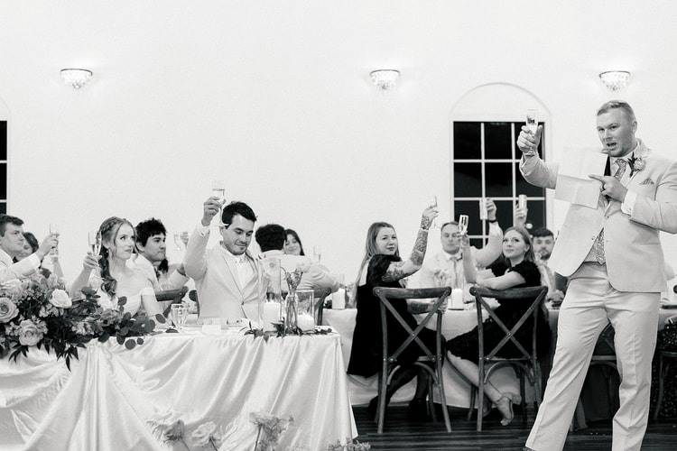 Black and white shot of the wedding party raising champagne glasses in a toast as a groomsman speaks into a microphone.