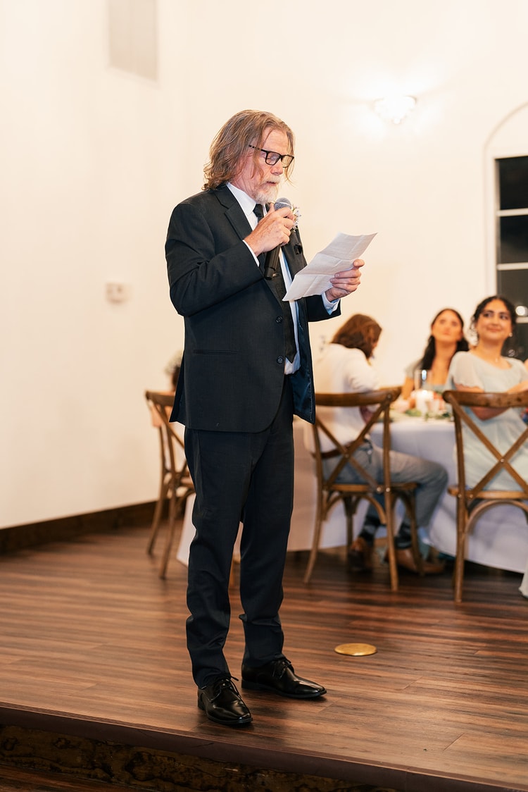 Black and white portrait of an older man with long hair and glasses giving a heartfelt speech into a microphone at the reception.