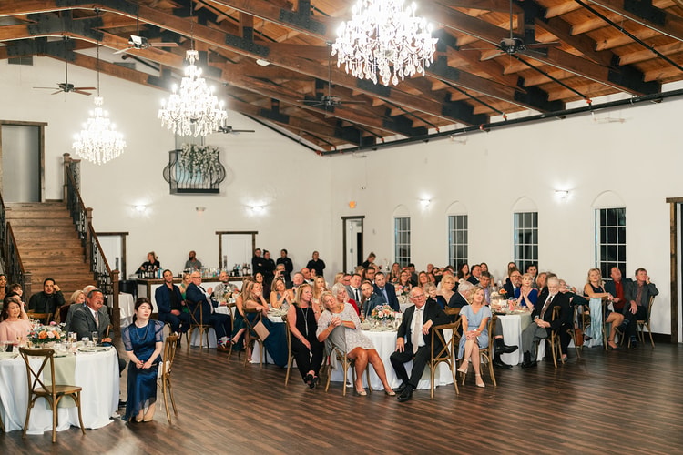 Wide interior view of the reception hall with guests seated at round tables beneath multiple crystal chandeliers and a wooden staircase.