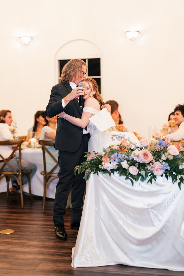 Father embraces the bride while speaking into a microphone beside the flower-draped head table during his wedding toast.