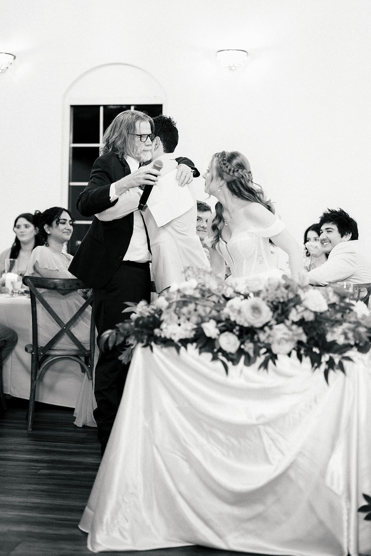 Black and white candid of a father hugging the groom while the bride laughs beside them during a reception toast.