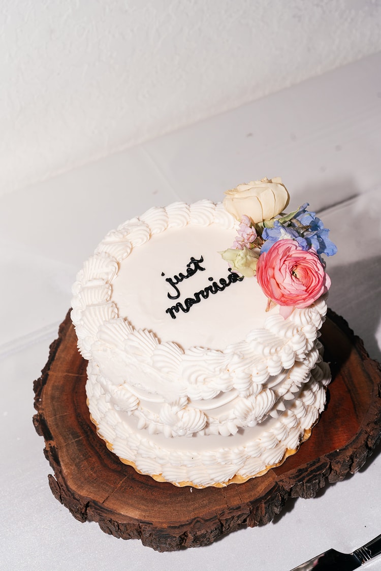 Small round wedding cake reading 'just married' in script, topped with fresh roses and ranunculus, set on a rustic wood slab.