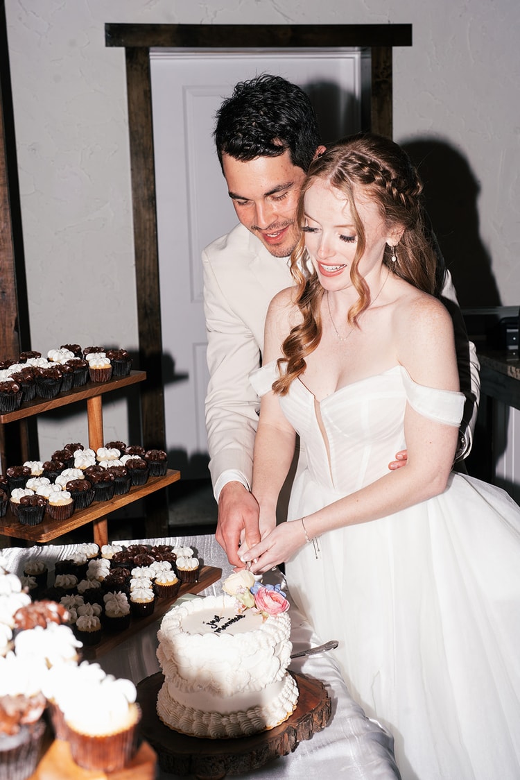 Bride and groom cut into their small tiered wedding cake together beside trays of chocolate and vanilla cupcakes.