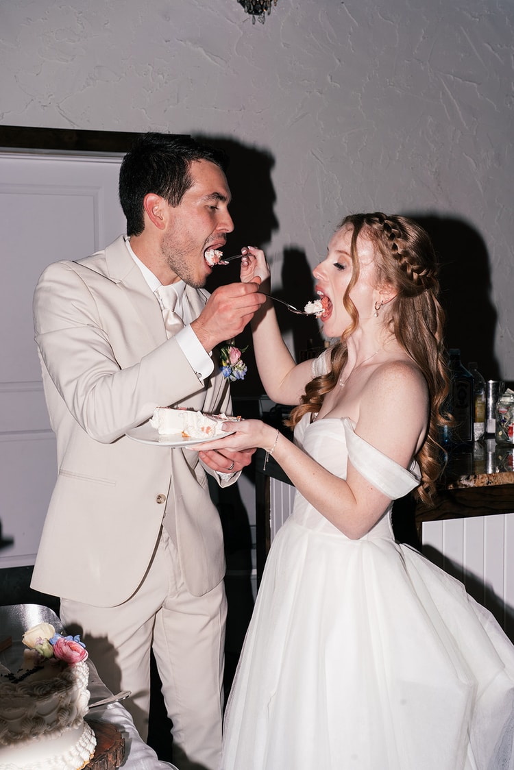 Bride and groom feed each other bites of wedding cake, both laughing mid-bite at the dessert table.