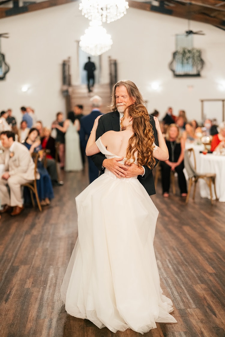 Father dances with his bride during their reception dance, guests seated at surrounding tables under chandeliers.