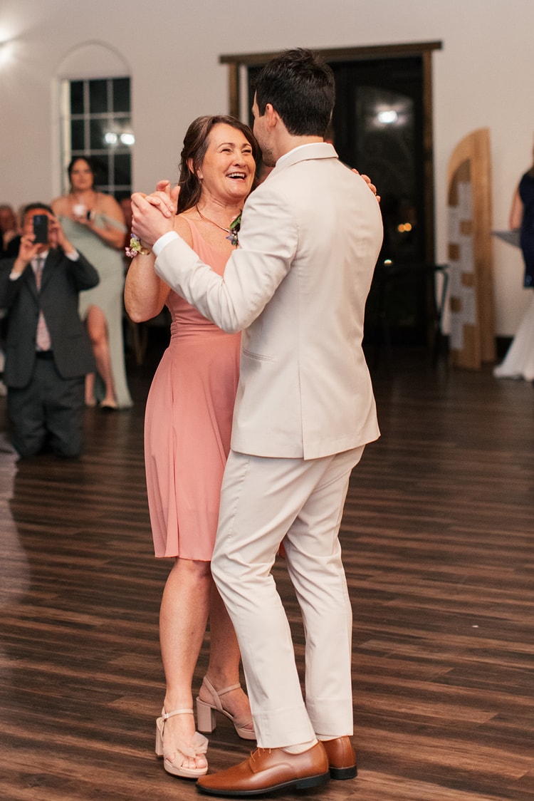 Groom dances with his mother in a pink dress on the reception floor, both smiling as guests photograph the moment.