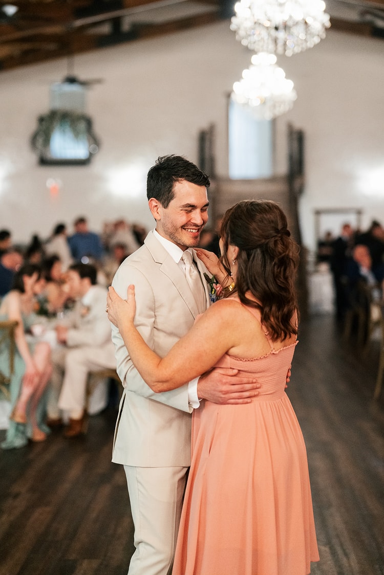 Groom laughs while dancing with a bridesmaid on the reception floor, string lights and chandeliers overhead.