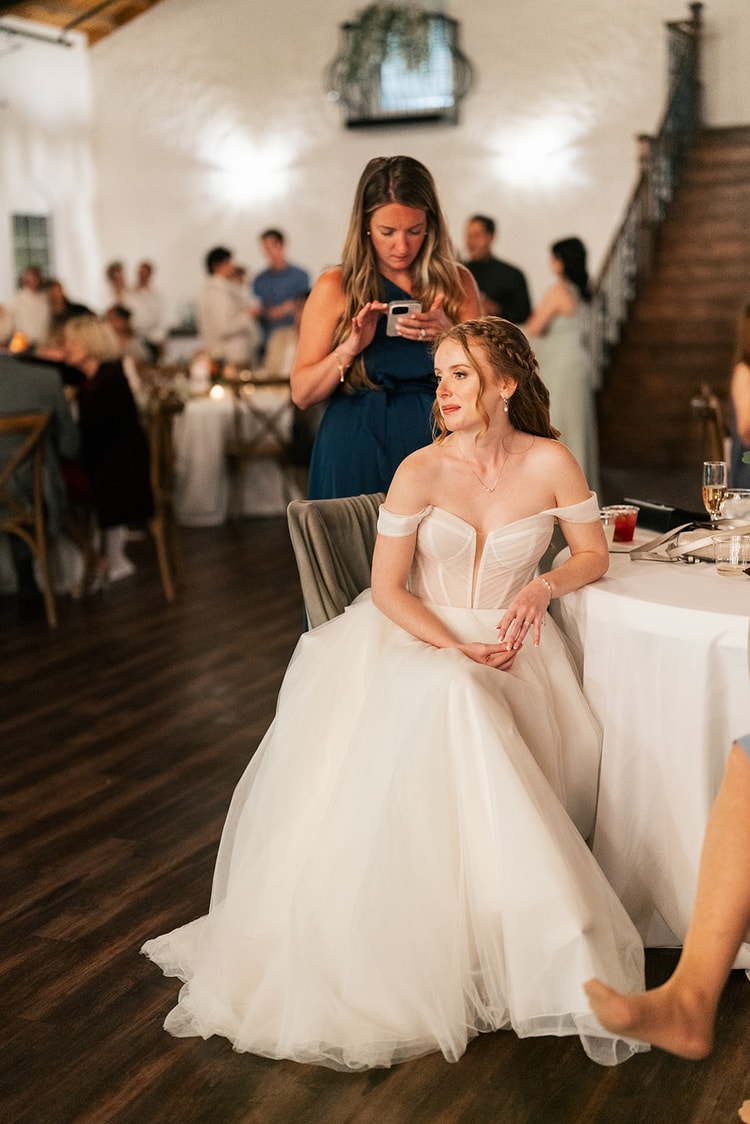 Bride sits at the sweetheart table watching the room while a guest photographs her, ballgown pooling on the floor.