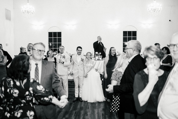 Wedding party stands together on the dance floor as a guest gives a toast into a microphone, black and white.