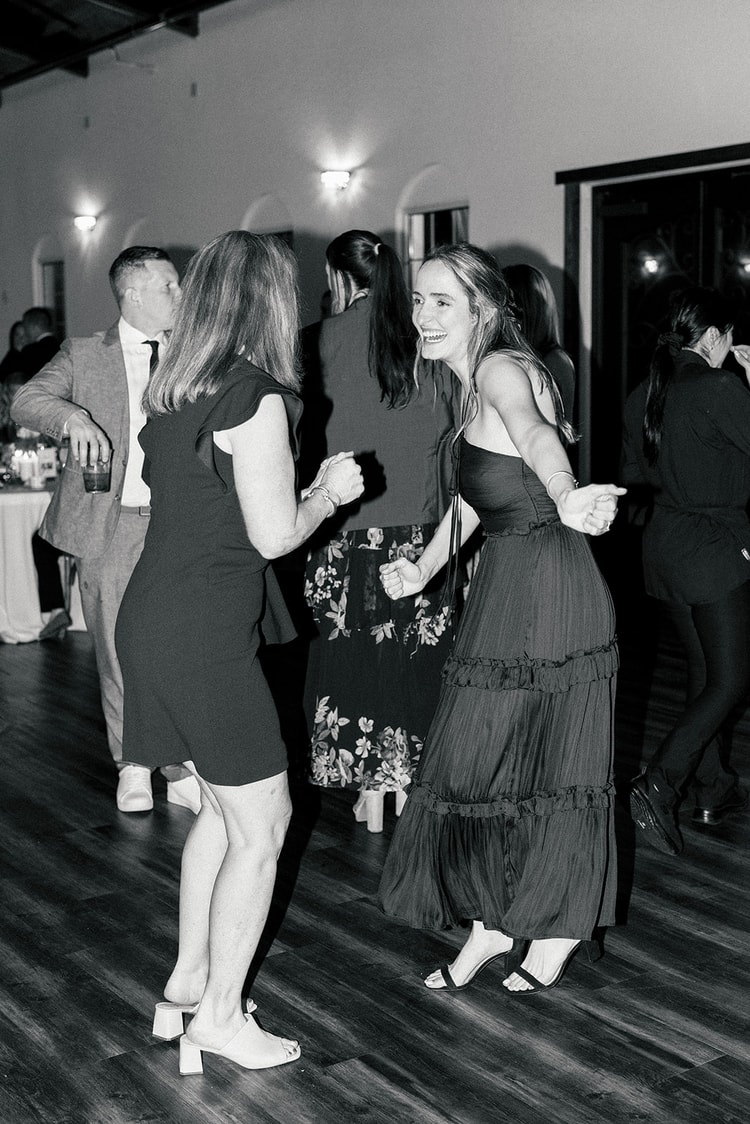 Wedding party and officiant stand together on the dance floor as guests gather around for a reception toast, black and white.