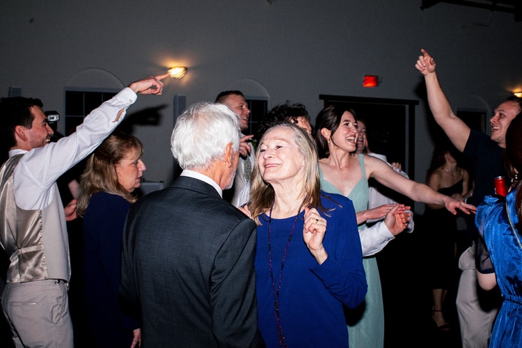 Guests link arms and cheer during a lively group dance at the reception, one man pointing toward the ceiling lights.