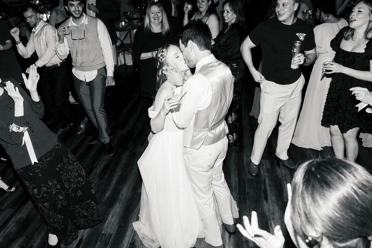 Bride and groom kiss while dancing surrounded by guests on a crowded dance floor, black and white.