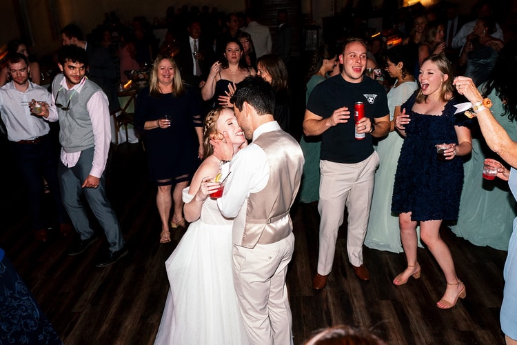 Bride and groom embrace and laugh on the dance floor while she holds a cocktail, surrounded by cheering guests.