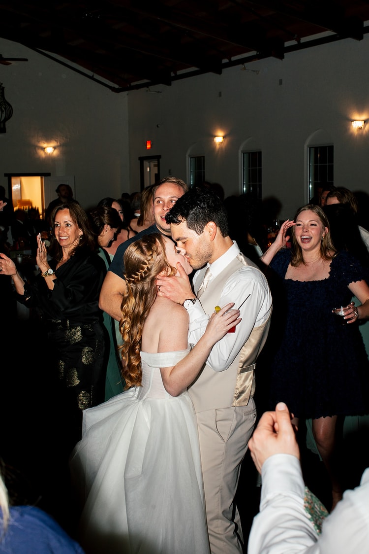 Bride and groom kiss on the dance floor as an officiant looks on, groom holding a drink, black and white.