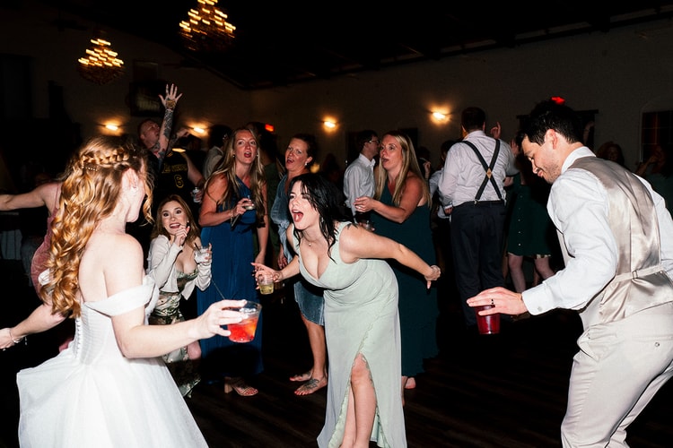 Bride dances in a circle of friends, one bridesmaid leaning back mid-laugh with a drink in hand at the reception.
