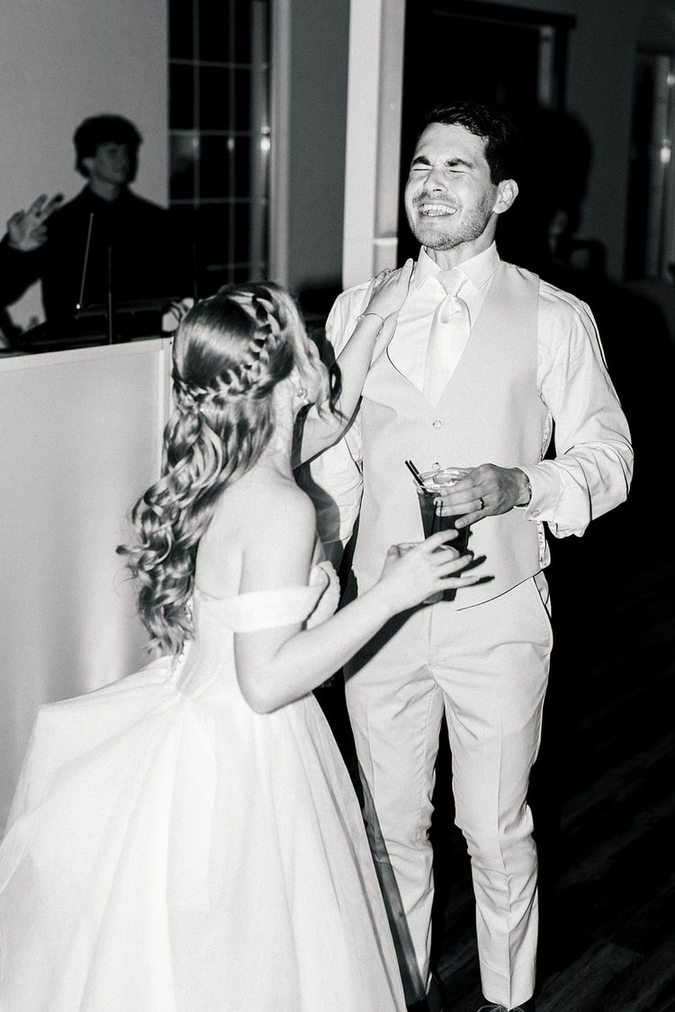 Bride laughs while holding the groom's arm and a drink as he smiles broadly, close moment on the dance floor.