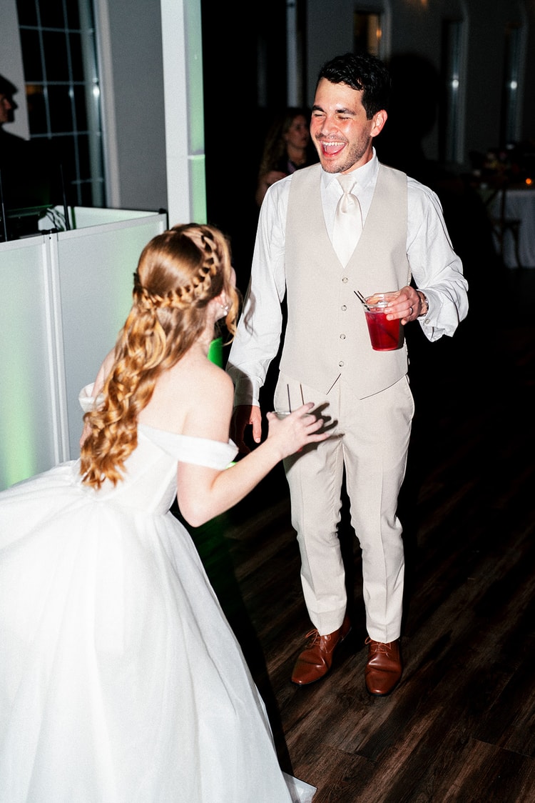 Bride reaches for her laughing groom's hand as they dance together at the Orlando reception, both holding drinks.