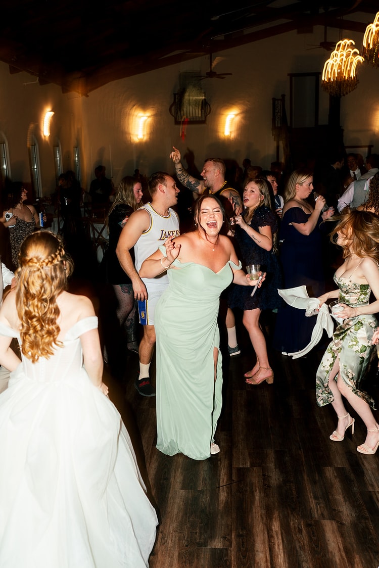 Bridesmaid in a sage green dress laughs and dances with the wedding party on a crowded reception floor.