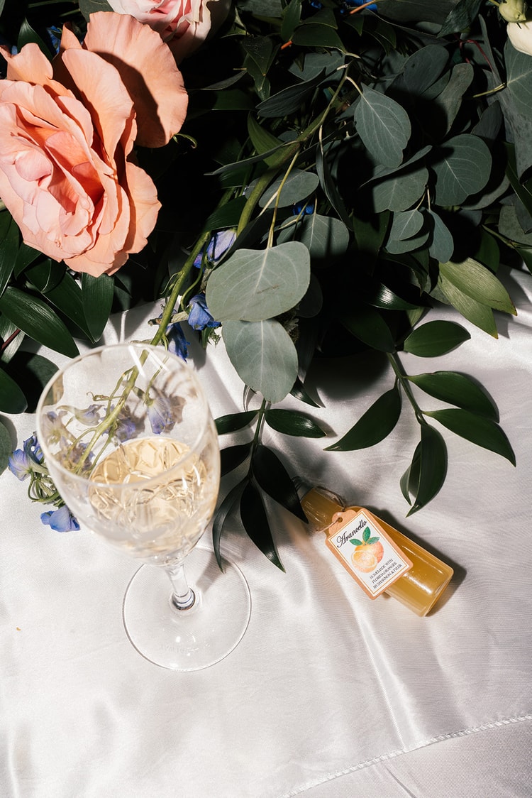 Overhead detail of a champagne glass, blush roses, eucalyptus, and a bottled limoncello favor on a satin tablecloth.