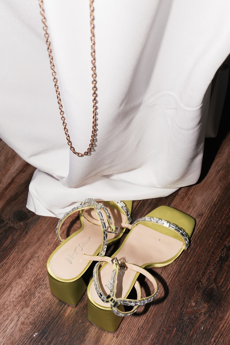 Bride's green strappy heels and a necklace rest on the wood floor beneath her gown during a reception break.