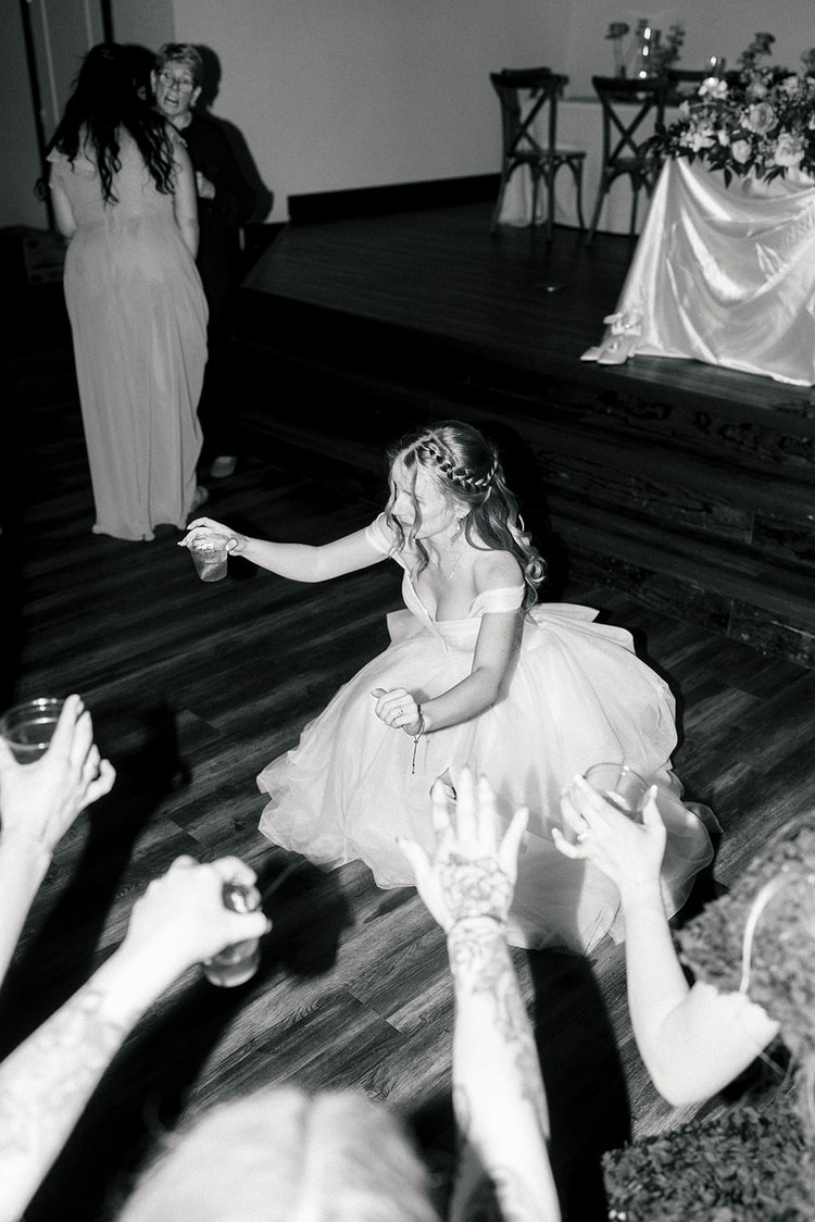 Bride dances near the stage, cup raised, as guests reach up around her, black and white low-light shot.