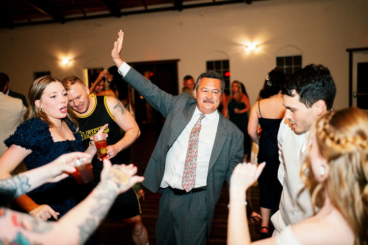 Guest in a gray suit raises his arm and dances joyfully in a circle of wedding guests at the reception.