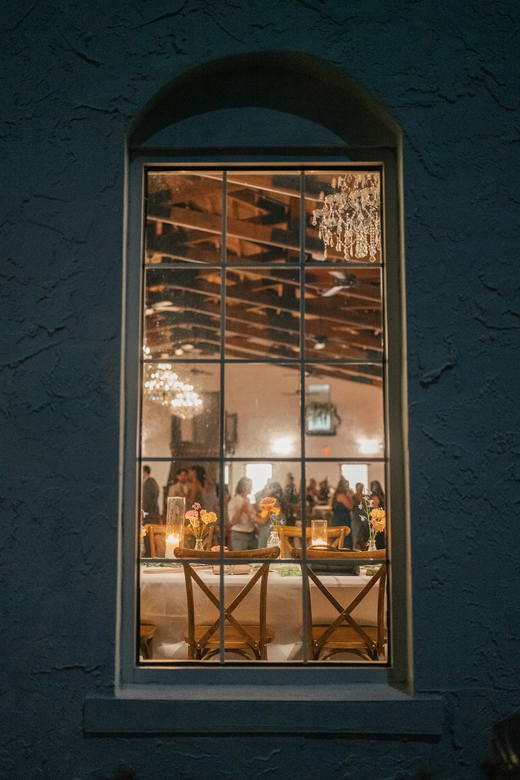 Arched window of the Orlando reception hall reflecting chandeliers and guests gathered around a floral-topped table at night.