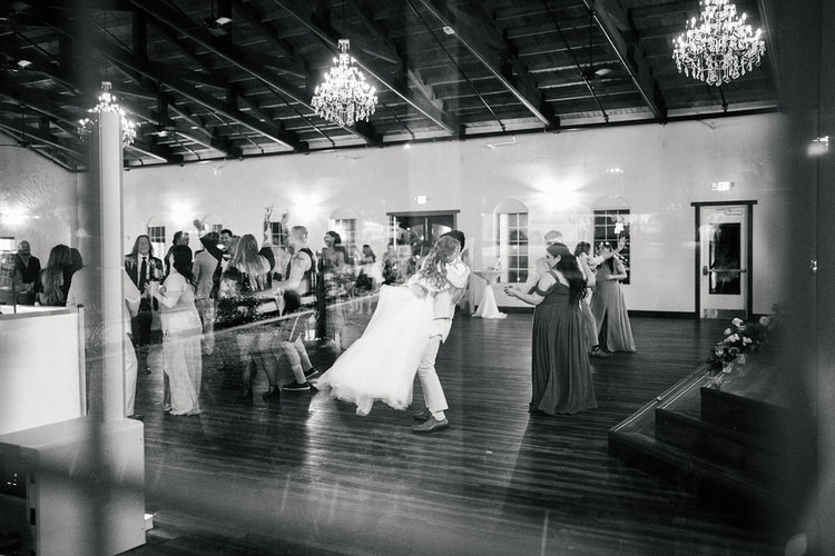 Groom dips and kisses the bride on the dance floor as guests dance nearby, chandeliers glowing above, black and white.