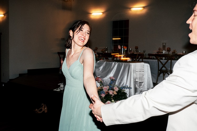 Bridesmaid in a sage dress laughs while holding the groom's hand mid-dance near the reception's floral sweetheart table.