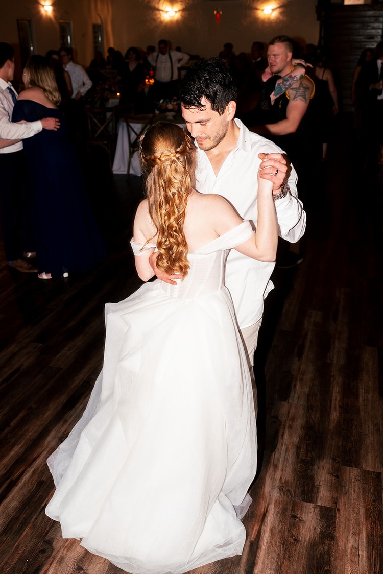Groom dances closely with his bride, her braided hair catching the light, guests visible in the background.