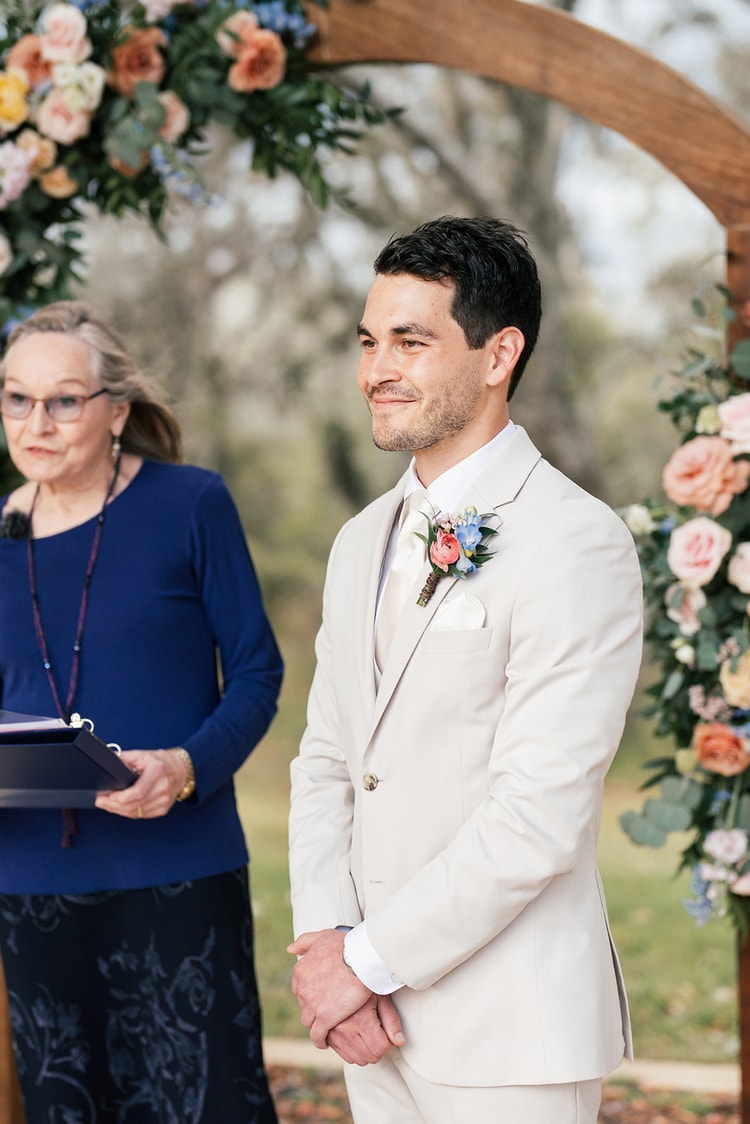 Groom in a cream suit and floral boutonniere smiles at his bride during the outdoor ceremony as the officiant looks on nearby.