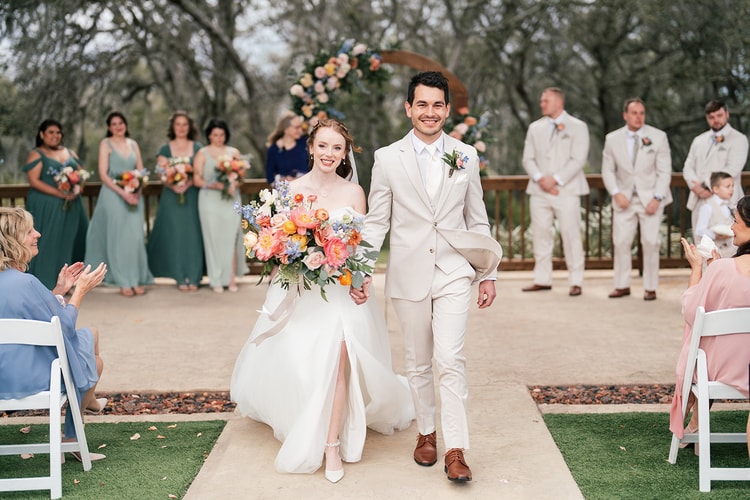 Newlyweds walk back up the aisle smiling and holding hands as guests applaud on either side.