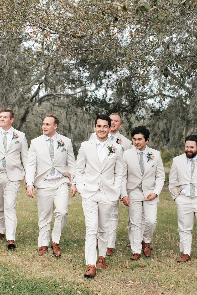 Groomsmen in cream suits walk together beneath a canopy of moss-covered oak branches, several smiling and looking off to the side.