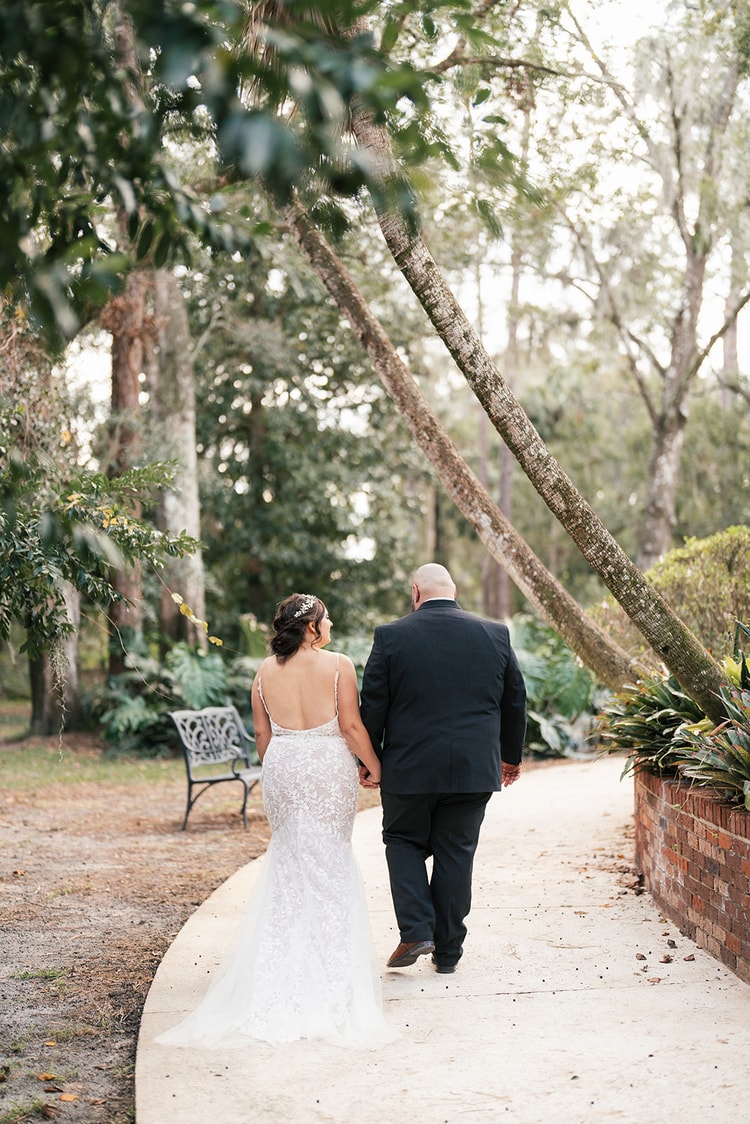 Bride and groom walking hand in hand away from the camera along a winding garden path, her lace train flowing behind them beneath moss-draped trees