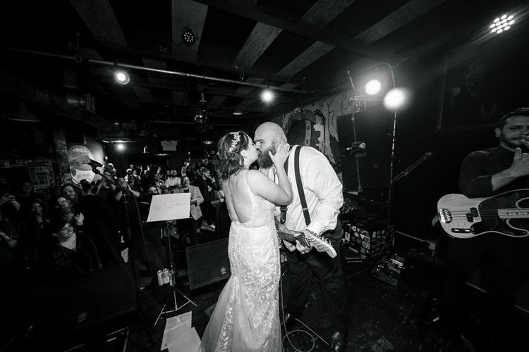 Black and white wide shot of the groom kissing the bride on stage at the music venue reception, guitar still in hand, as a full crowd of celebrating guests looks on