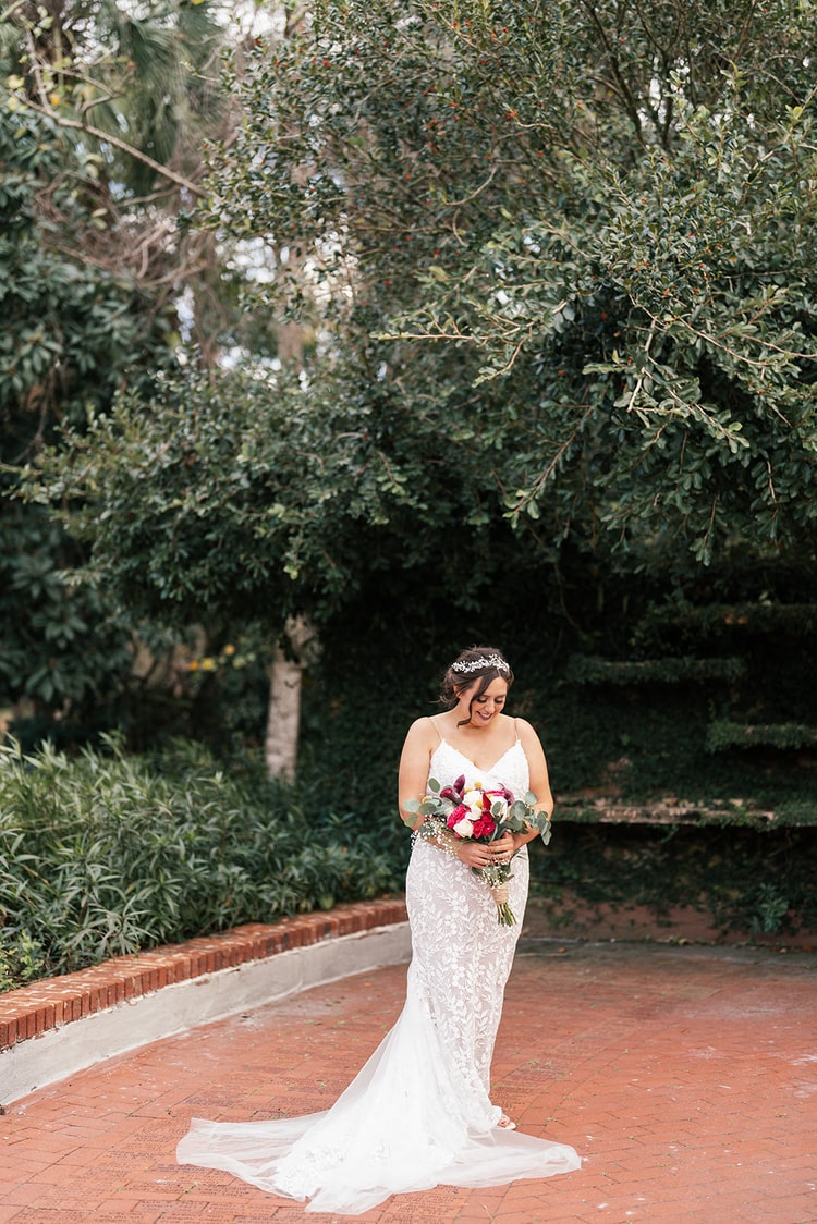Bride in a fitted lace gown and crystal hair vine standing alone on a brick courtyard, looking down at her bouquet with lush green hedges and trees behind her