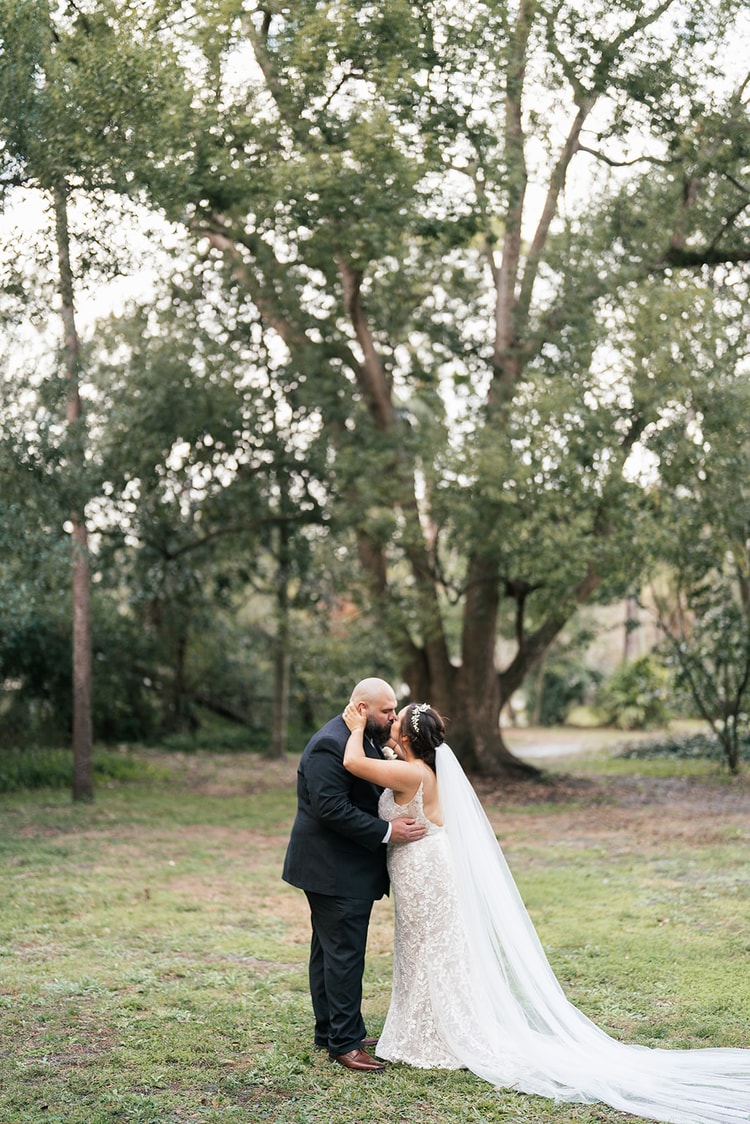 Bride and groom sharing their first kiss after the ceremony, embracing beneath a white arbor with flowering shrubs and trees in the background